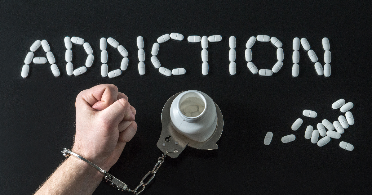 The word “addiction” spelled out in pills beside a handcuffed hand and an overturned pill bottle.